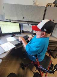 BH-PHASE-web Physically disabled man in blue shirt and baseball cap sits on a walker and types at a computer station