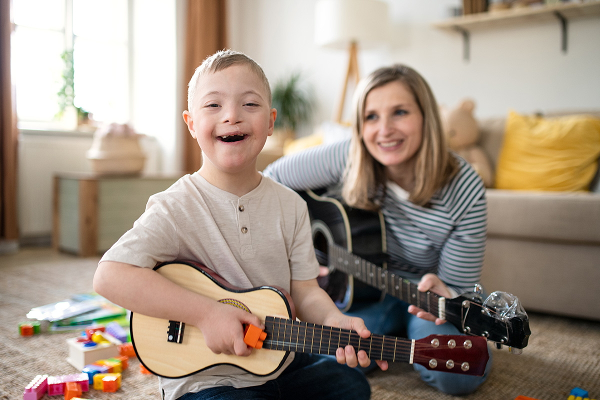 Boy with Down's Syndrome sits on the floor in a living room next to a blonde woman. Both are smiling. The boy is playing a ukelele and the woman is playing a guitar.