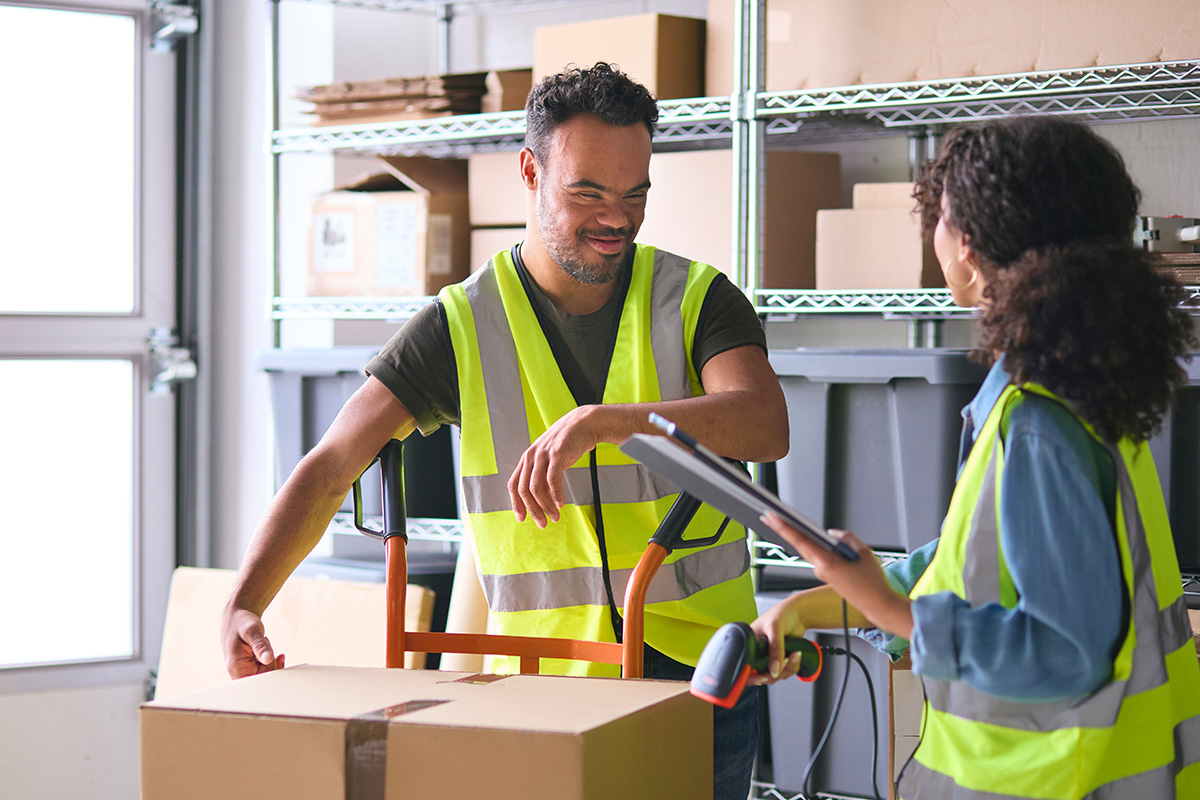 Woman Using Digital Tablet And Male Colleague With Down Syndrome Working In Warehouse