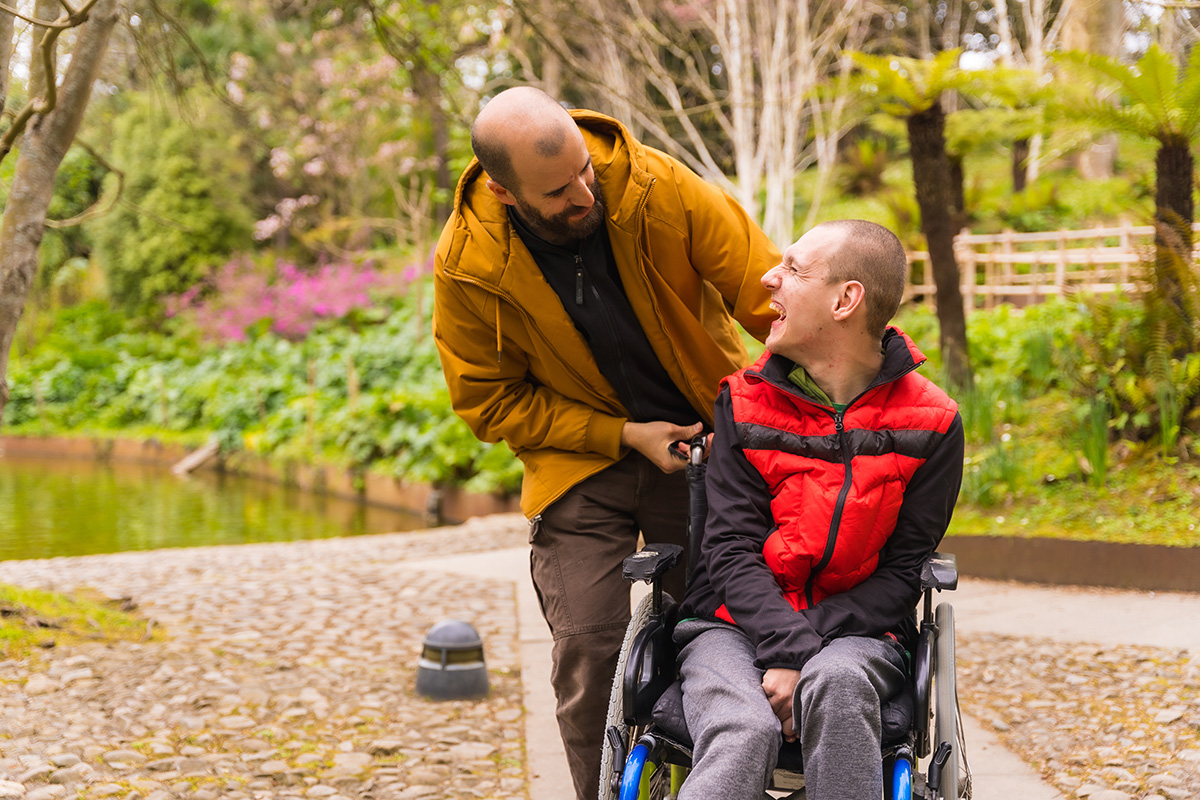 A middle aged male caregiver pushed a disabled young adult male through a lakeside park in a wheelchair. The caregiver is wearing a winter coat and the young man is wearing grey slacks and a red vest. The two men are talking and smiling.