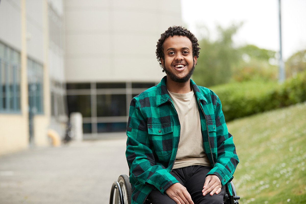 Somali man in a wheel chair sits outside a building next to a small hill. He is wearing a T-shirt and a green plaid flannel shirt. He is smiling.