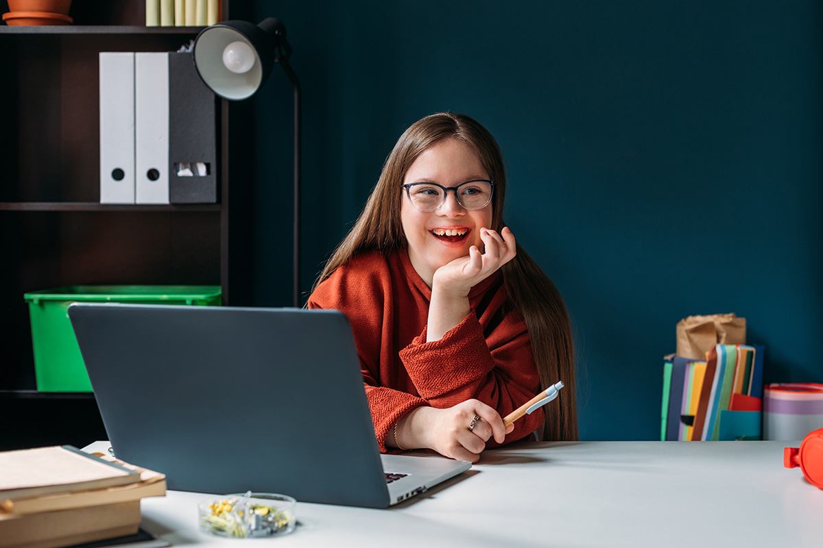 Woman sits at a desk in an office with dark walls and book shelves behind her. She sitting in front of an open laptop, holding a pen and smiling.