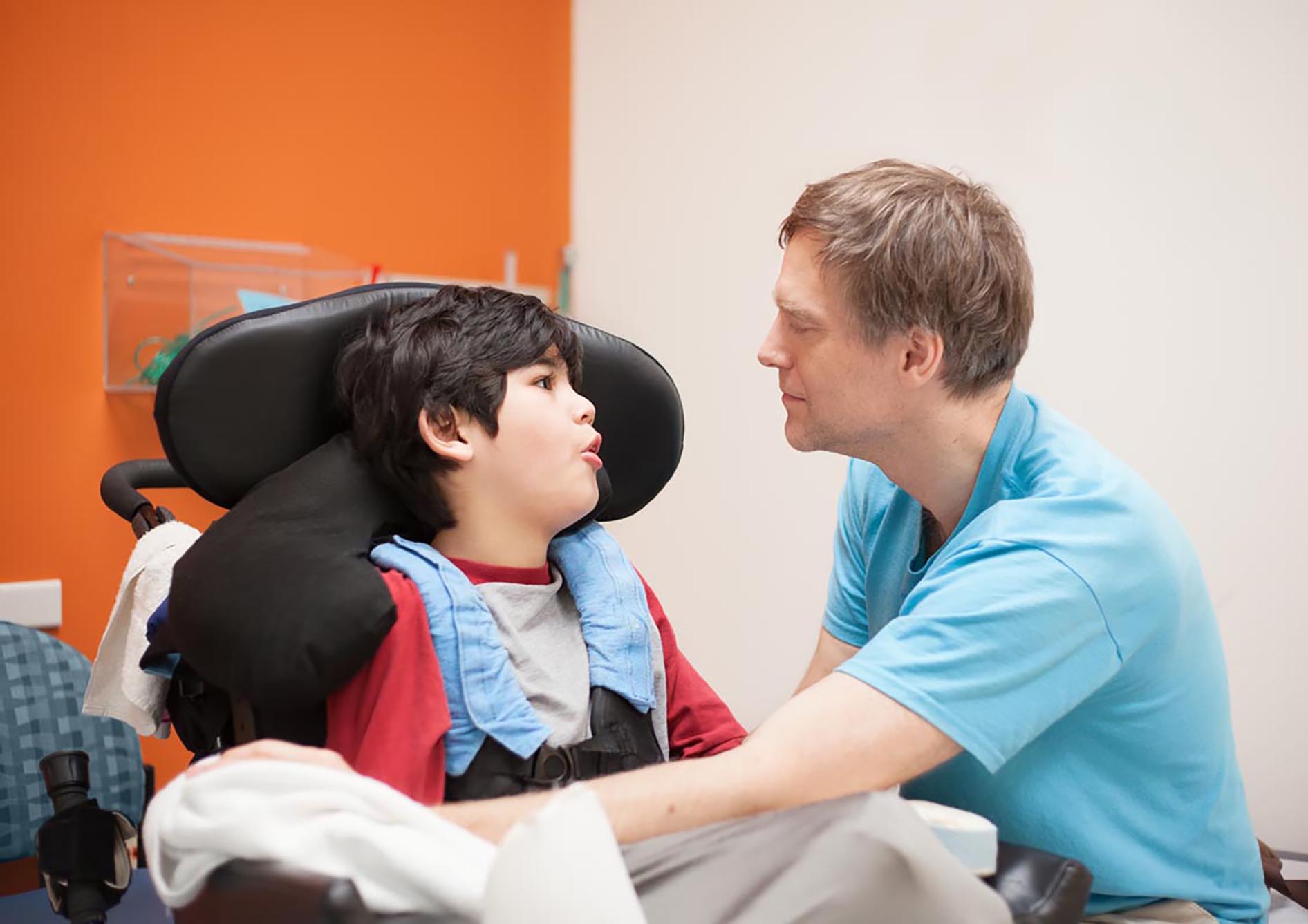 male caregiver in hospital scrubs crouches down to help young boy in a wheelchair