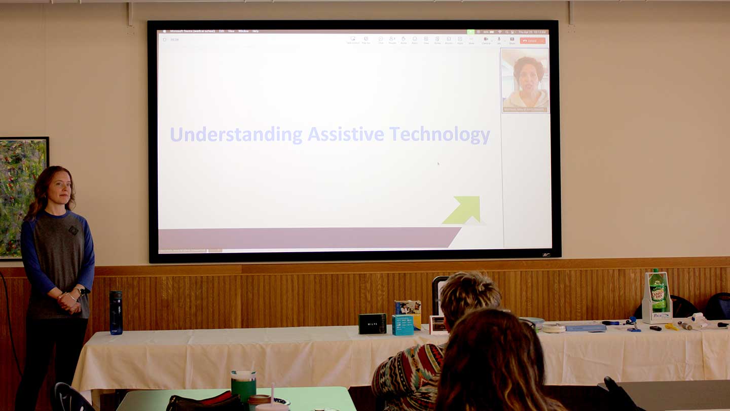 A presenter, a blond woman in her 30s wearing a gray sweater and black slacks, stands om front of a screen that reads Understanding Assistive Technology