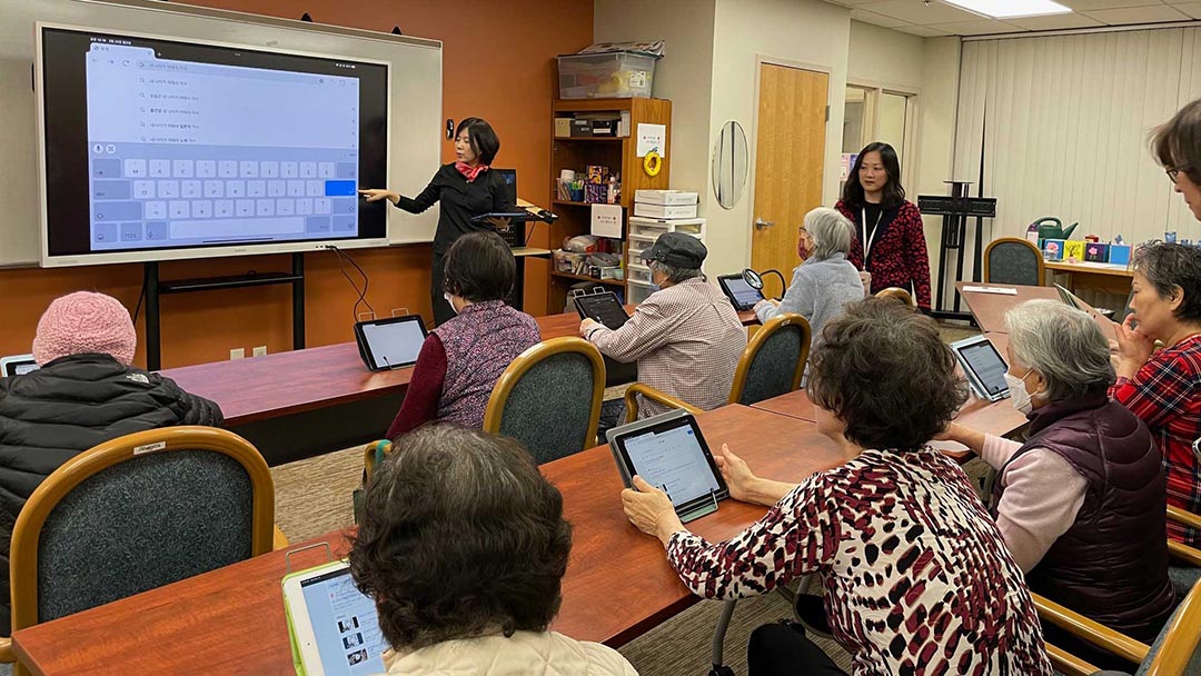 A middle aged woman with dark hair and wearing a black dress and red scarf, makes a presentation about how to use an iPad to Korean elders sitting in a conference room. The presenter points to an image of a keyboard on a projector screen