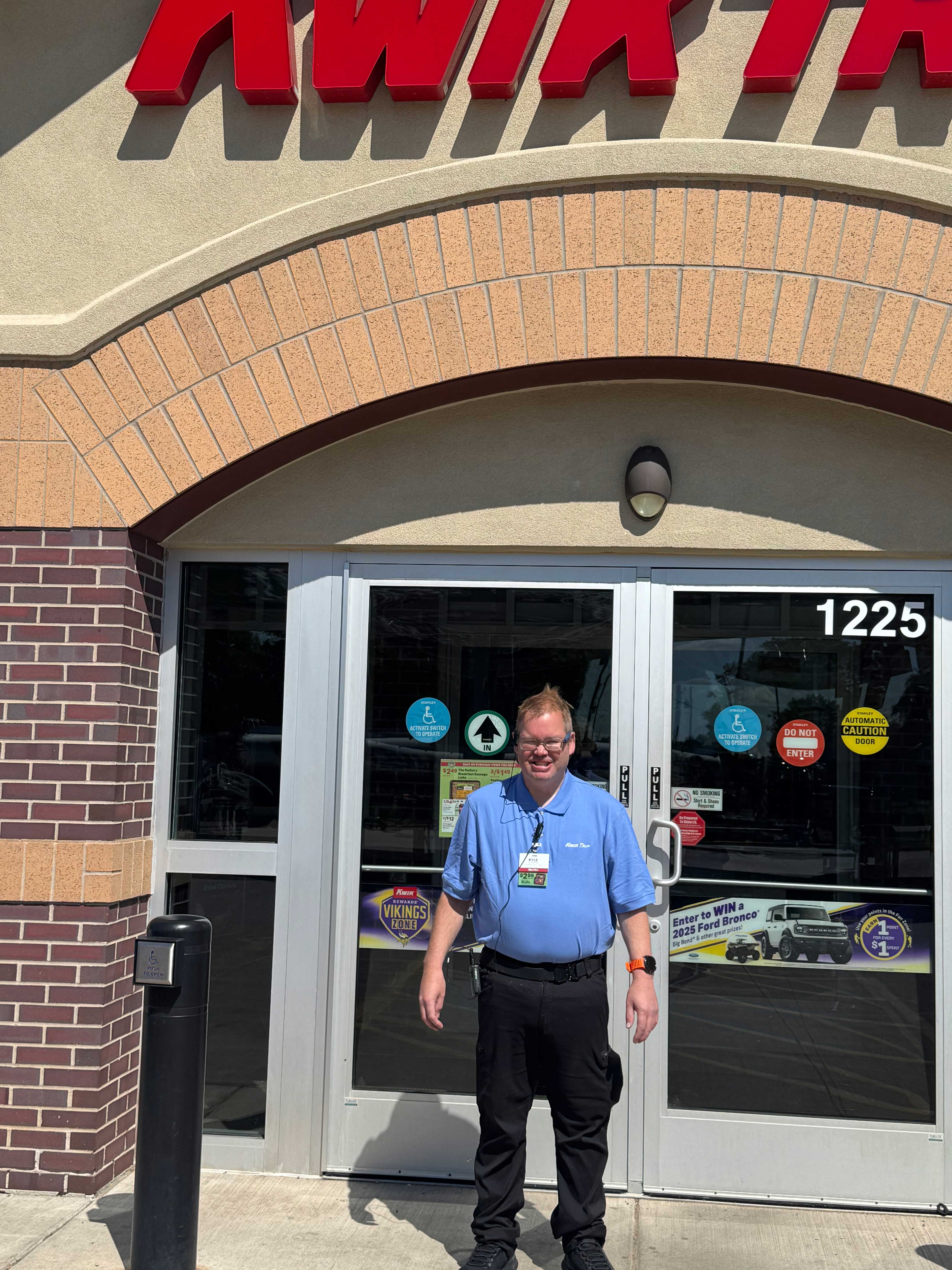 Man in blue polo shirt and black pants, wearing a work badge on a lanyard, stands in front of a Kwik Trip convenience store