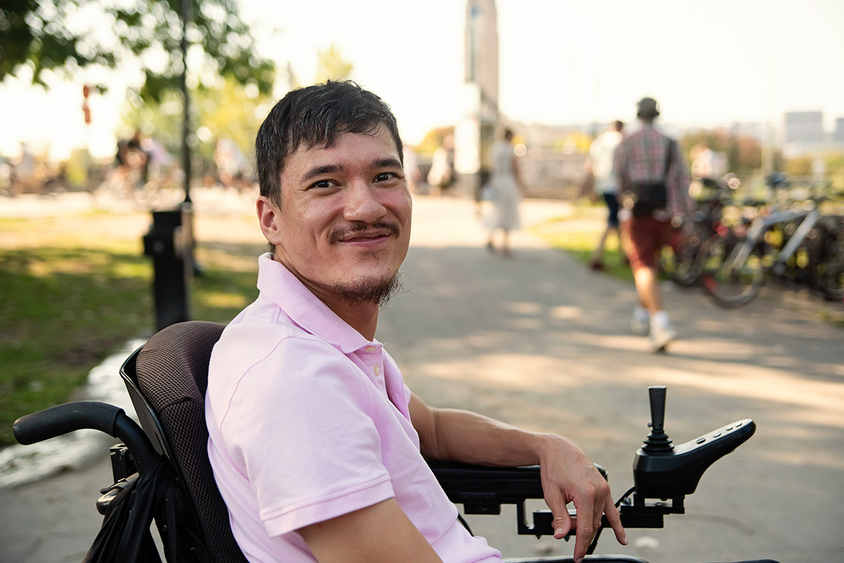 Asian man with short black hair and mustache sits in a wheelchair in a park. He is wearing a pink polo shirt