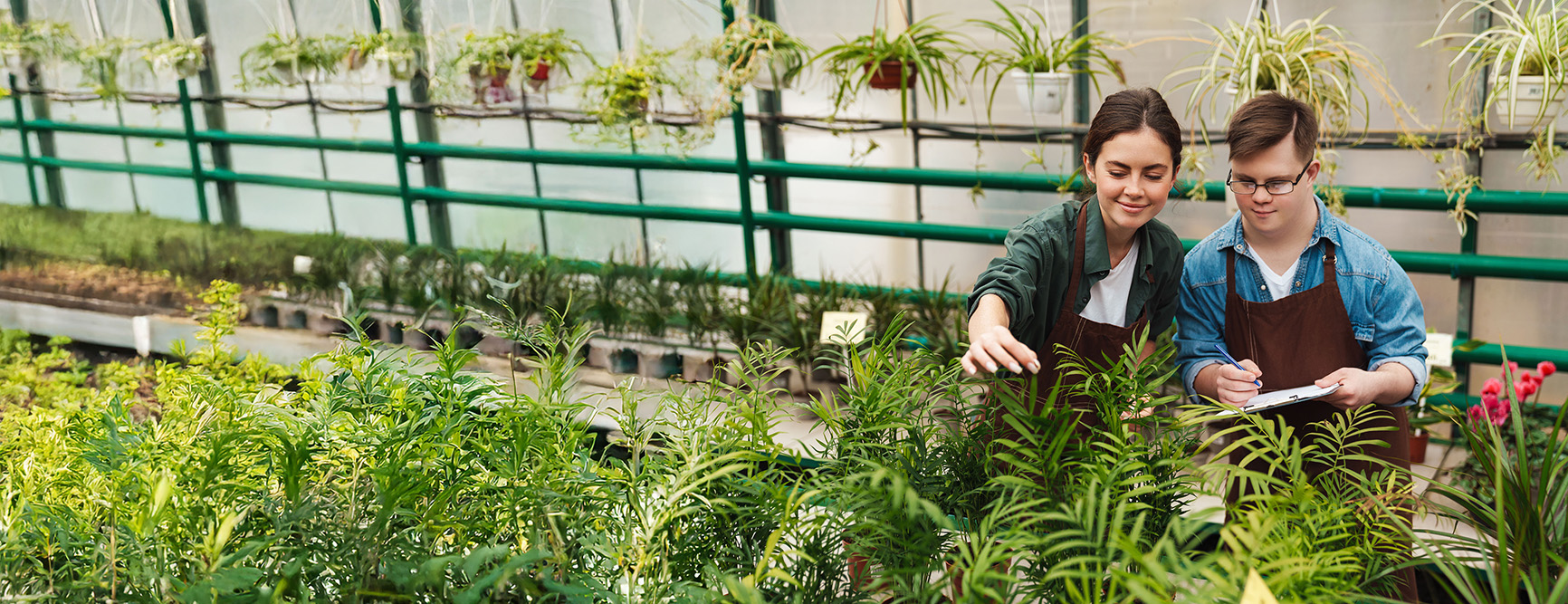 A young woman with dark hair pulled back, wearing a green flannel shirt, white t-shirt and brown apron helps a young man with Down's Syndrome wearing a denim shirt, white t-shirt and brown apron. They are working in a plant-filled greenhouse. The young man uses a pen to take notes on a clipboard. 